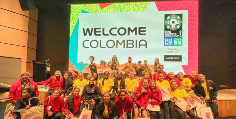 Mujeres de la Selección Colombia femenina, usando chaquetas amarillas y rojas, posan reunidas frente a un letrero que dice 'Bienvenido Colombia'