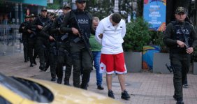Policías armados custodiando a un hombre con hoodie blanco y shorts rojos